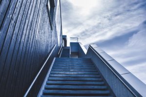 A modern, blue-toned outdoor staircase in Washington D.C. ascends between two building walls under a partly cloudy sky. The perspective creates a dynamic, upward view, highlighting the architectural design and contrasting textures of the wood and concrete surfaces while ensuring safety.