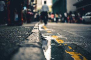 A city street scene features a wet pavement and yellow lines, with a shallow depth of field. People in the background are blurred, suggesting movement and a bustling urban environment. Nearby, an injured pedestrian sits on the sidewalk while a puddle reflects towering buildings above.