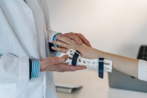 In a bright Washington D.C. clinic, a doctor in a white coat examines a patient's forearm fitted with an orthopedic splint secured by blue straps. As part of the Workers' Compensation process, the doctor gently holds the patient's hand, with a computer visible in the background.