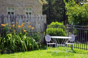 A cozy backyard setting with a metal table and two matching chairs on a grassy lawn. Bright orange flowers and lush greenery border the area, where guests can relax without worrying about injury liability. A stone house and wooden fence provide a charming backdrop.