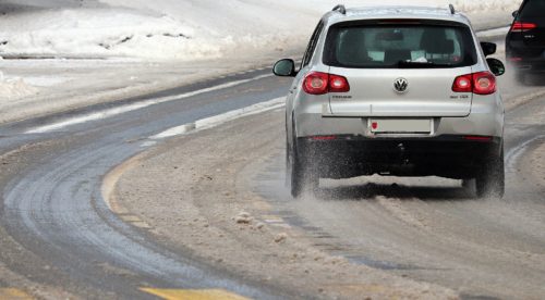 A white SUV drives cautiously along a snow-covered, wet road with tire tracks visible. Snow is piled on the roadside, and another vehicle is seen ahead. The scene implies winter conditions and highlights the responsibility for passenger injury in such treacherous driving conditions.
