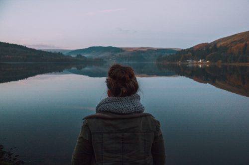 A person with a bun hairstyle and a scarf stands by a calm lake, recently recovered from an accident case. Surrounded by hills under a pastel sky at dusk, the scene reflects on the water, creating a mirror-like effect.