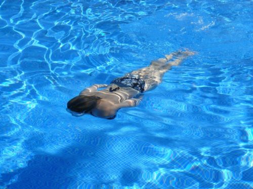 A woman swims underwater in a clear blue pool, wearing a black bikini with a floral pattern. As sunlight creates ripples on the water's surface, adding to the serene atmosphere, remember pool safety tips to ensure your Labor Day weekend remains as calm and beautiful as this moment.