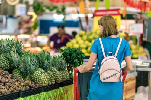 A woman in a blue dress with a pink backpack shops for fruit in a vibrant Washington D.C. market. She looks at a display of pineapples and other tropical fruits, while the background shows more produce and shoppers bustling through the aisles.