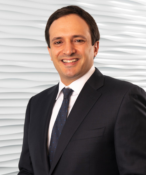 A smiling man in a dark suit and tie, Daniel S. Singer, stands confidently in front of a white textured wall.