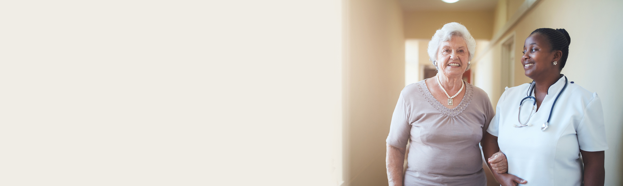 A senior woman in a light purple top walks arm-in-arm with a healthcare professional in a white uniform and stethoscope down a well-lit corridor, both smiling. washington D.C nursing home abuse lawyer concept image.