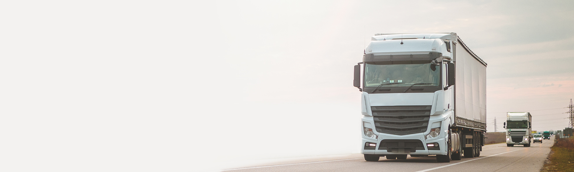 Two large white trucks with trailers driving on a highway, heading towards the camera on a clear day. The background features an expansive sky and a wide open road.