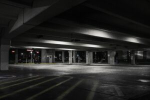 A dimly lit, empty parking lot with concrete pillars and beams casts an eerie aura. Yellow markings stretch across the floor, while small lights provide faint illumination. Outside, blurred trees and distant streetlights peer through the openings like ghosts haunting the night.