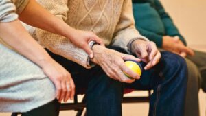 A close-up of a seated elderly person holding a colorful stress ball, wrapped in a knitted sweater. Another individual, in a gray outfit, gently supports their arm, offering comfort. A third person is partially visible beside them, highlighting the importance of vigilance against nursing home abuse.