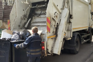 A sanitation worker in a dark uniform is briskly loading black garbage bags into the back of a white garbage truck adorned with red and yellow reflective stripes, ensuring that nothing will hit the garbage truck as it moves along the street.