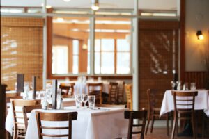 A quaint restaurant interior with wooden chairs and tables covered in white tablecloths. Glasses and cutlery are neatly arranged on the tables, ready to sue any culinary challenge. Large windows with bamboo shades allow natural light to fill the space beautifully.