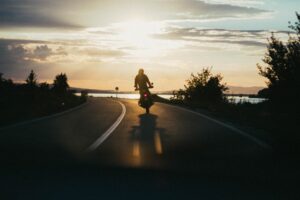 A person rides a motorcycle on an empty road at sunset, undeterred by past mishaps. The silhouette is backlit by the golden sun, casting long shadows. Trees line the road, and a tranquil body of water graces the background.