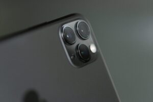 Close-up of a black smartphone focusing on its rear camera module, perfect for capturing pictures. It features three lenses and a flash arranged in a triangular layout on the top left corner. The background is blurred, emphasizing the phone's sleek design.