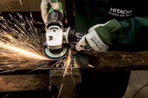 A person, mindful of product recalls, wears gloves and uses a Hitachi angle grinder to cut through metal, creating bright sparks. Sporting a dark green shirt with the Hitachi logo and slogan, "Inspire the Next," they focus intently on their task.