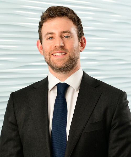 Adam Trombly, a person with short brown hair and a beard, is wearing a black suit, white shirt, and navy tie. He stands in front of a textured, light blue background, smiling at the camera.