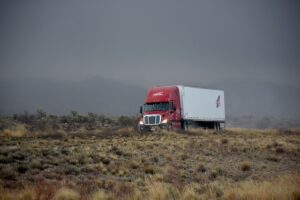 A red and white semi-truck navigates a rough, grassy landscape under an overcast sky, with mountains faintly visible in the background. The scene conveys a sense of isolation and ruggedness, as if the truck itself is defying the elements in its steadfast journey.