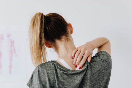 A woman with a ponytail faces away from the camera, wearing a green shirt. Her hand rests on her neck, gently massaging the area for pain management. The background features a white wall with a blurry human body chart image.