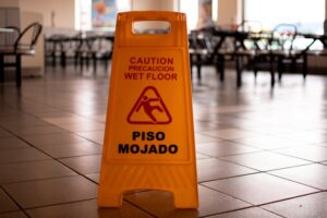 A yellow caution sign on a tiled floor reads "Caution: Wet Floor" in English and Spanish ("Precaución: Piso Mojado"). The background features a blurry empty restaurant with chairs and tables.
