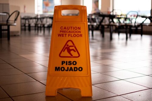 A yellow caution sign on a tiled floor reads "Caution: Wet Floor" in English and Spanish ("Precaución: Piso Mojado"). The background features a blurry empty restaurant with chairs and tables.