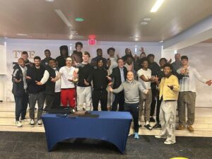 Ken Trombly and the GW Men's Basketball Team pose indoors for a photo. Some are dressed in athletic wear while others opt for business casual. A table with a blue tablecloth stands before them, adding charm to the scene as the partially visible sign "THE" entertains with curious mystery.