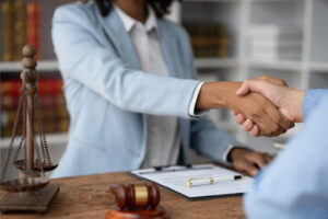 Two people shaking hands across a desk in an office, with legal documents, a judge's gavel, and scales of justice visible, suggesting an initial consultation with a personal injury attorney.