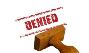 A wooden rubber stamp boldly emblazoned with the word "DENIED" in striking red letters casts its imprint on a white background, symbolizing firm rejection or refusal.
