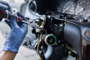 A person wearing blue gloves uses a screwdriver to repair or disassemble the steering column inside a car, with dashboard instruments, exposed wiring, and black box data components visible.