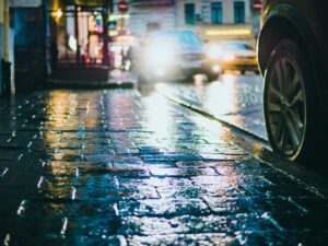 Wet cobblestone streets glisten with colorful neon lights, while blurred cars stream by in the background. A vehicle's wheel looms on the right, hinting at sidewalk hazards in this busy urban scene under a vibrant nighttime ambiance.