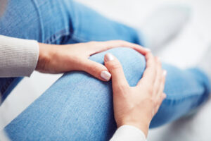 A person wearing blue jeans sits and holds their knee with both hands, suggesting possible knee pain or discomfort that may lead to a loss of enjoyment in life. The background is blurred, focusing attention on the hands and knee.