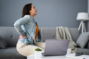 A woman with injuries sits on a gray sofa, holding her lower back in pain. Next to an open laptop on the coffee table, she worries about lost wages and can't work in her modern living room.