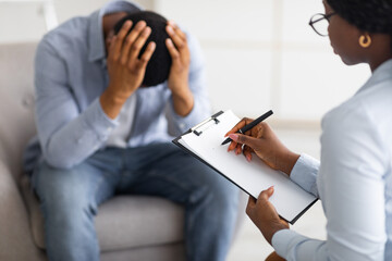 A person sits on a chair with their head in their hands, appearing distressed, possibly coping with PTSD, while a therapist nearby writes notes on a clipboard to help them recover.