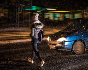 A person wearing winter clothes crosses a snowy street at night as a blue car approaches, its headlights shining—reminding us how quickly injuries can happen to pedestrians. The background glows with blurred lights from passing vehicles or buildings.