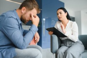 A woman holding a clipboard talks to a man who appears distressed, possibly discussing compensation for psychological injuries during a counseling session indoors.