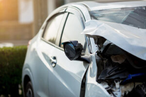 A close-up of a silver car with significant property damage to the front right side, including a crumpled fender and broken side mirror, parked near greenery in daylight after a car accident in Washington D.C.