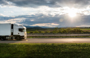 A white semi-truck from a reputable trucking company drives on a highway beside green fields, with distant hills and a bright sun shining through clouds in the sky.