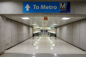 A hallway with gray walls and a shiny floor leads to a moving walkway; a blue sign above points straight ahead and reads "To Metro" with the Metro logo.
