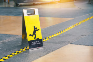 A yellow caution sign with a graphic of a person tripping and the text "BEWARE OF UNEVEN FLOOR" stands next to a floor marked with black and yellow tape, indicating a hazard in a public area.