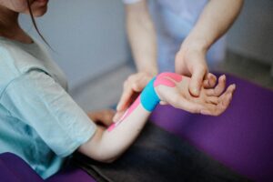 A person is having their forearm and wrist wrapped with colorful kinesiology tape by a healthcare professional in a clinical setting.