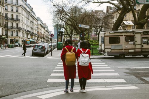 Two people wearing matching red coats and backpacks stand side by side at a crosswalk in a European city, with buildings, cars, and a parked camper van visible in the background.