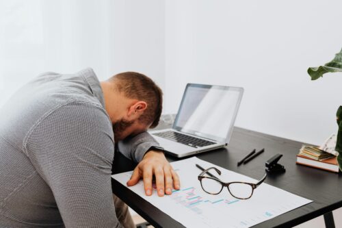 A man rests his head on his arm at a desk with a laptop, documents, glasses, and pens, appearing tired or stressed. A potted plant and books are also on the desk.