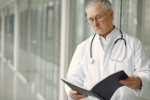 An elderly male doctor with gray hair and glasses, wearing a white coat and stethoscope, stands in a hallway reading from a black notebook—reflecting on the challenges of preventing medical misdiagnosis in Washington DC.