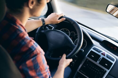 A person wearing a plaid shirt is driving a car, holding the steering wheel with both hands. The photo, taken in Washington D.C., captures the driver and dashboard from the passenger’s view, where head-on collisions are among common causes of accidents.
