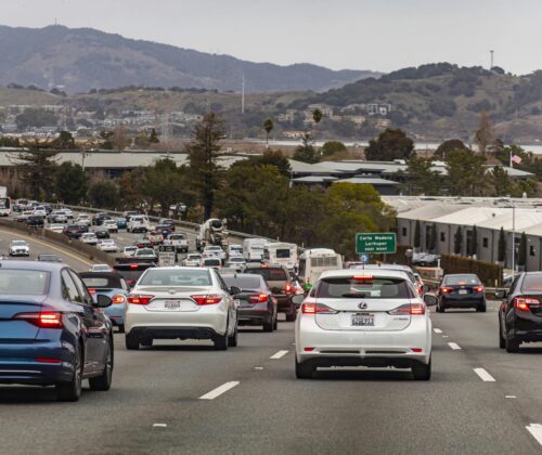 Heavy traffic on a multi-lane highway with cars closely packed, highlighting the importance of road safety. Mountains and trees are visible in the background under a cloudy sky, with a green exit sign on the right side of the road.