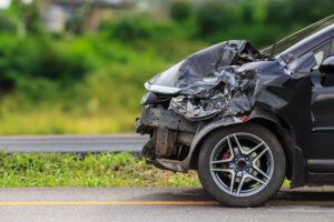 A black car with severe front-end damage sits on the side of a road in a green, blurred rural setting—an example highlighting factors affecting car accident value in Washington D.C.