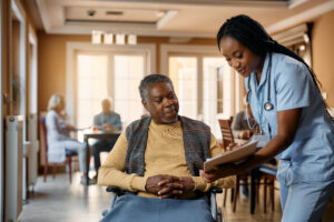 A nurse in scrubs smiles while showing a clipboard to an elderly man in a wheelchair. They are in a bright, communal room with other seniors and caregivers in the background.